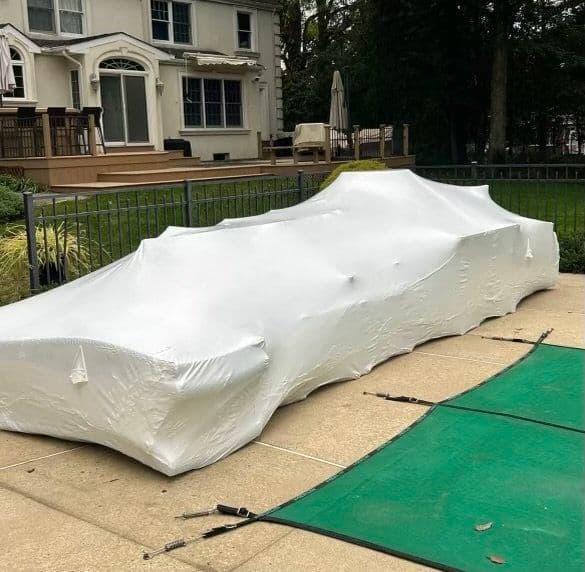 Car covered with white protective tarp in residential backyard next to deck and patio.