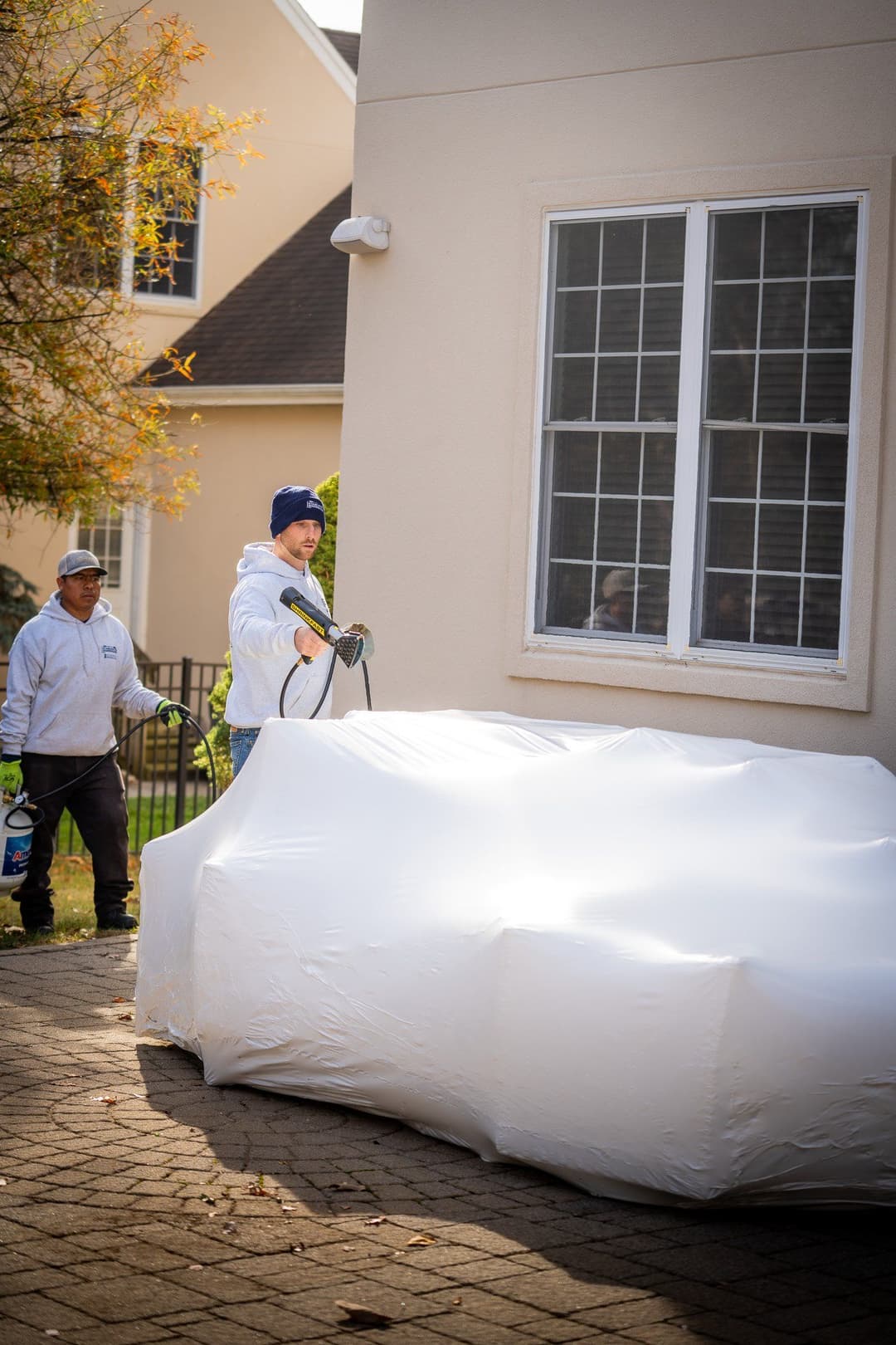 Workers applying protective cover to outdoor furniture near a house on a sunny day.