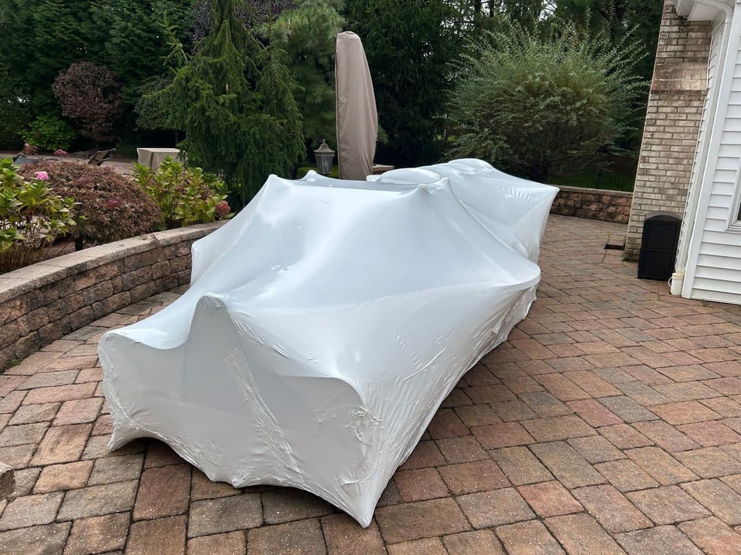 Outdoor furniture covered in white plastic on a brick patio with greenery in the background.
