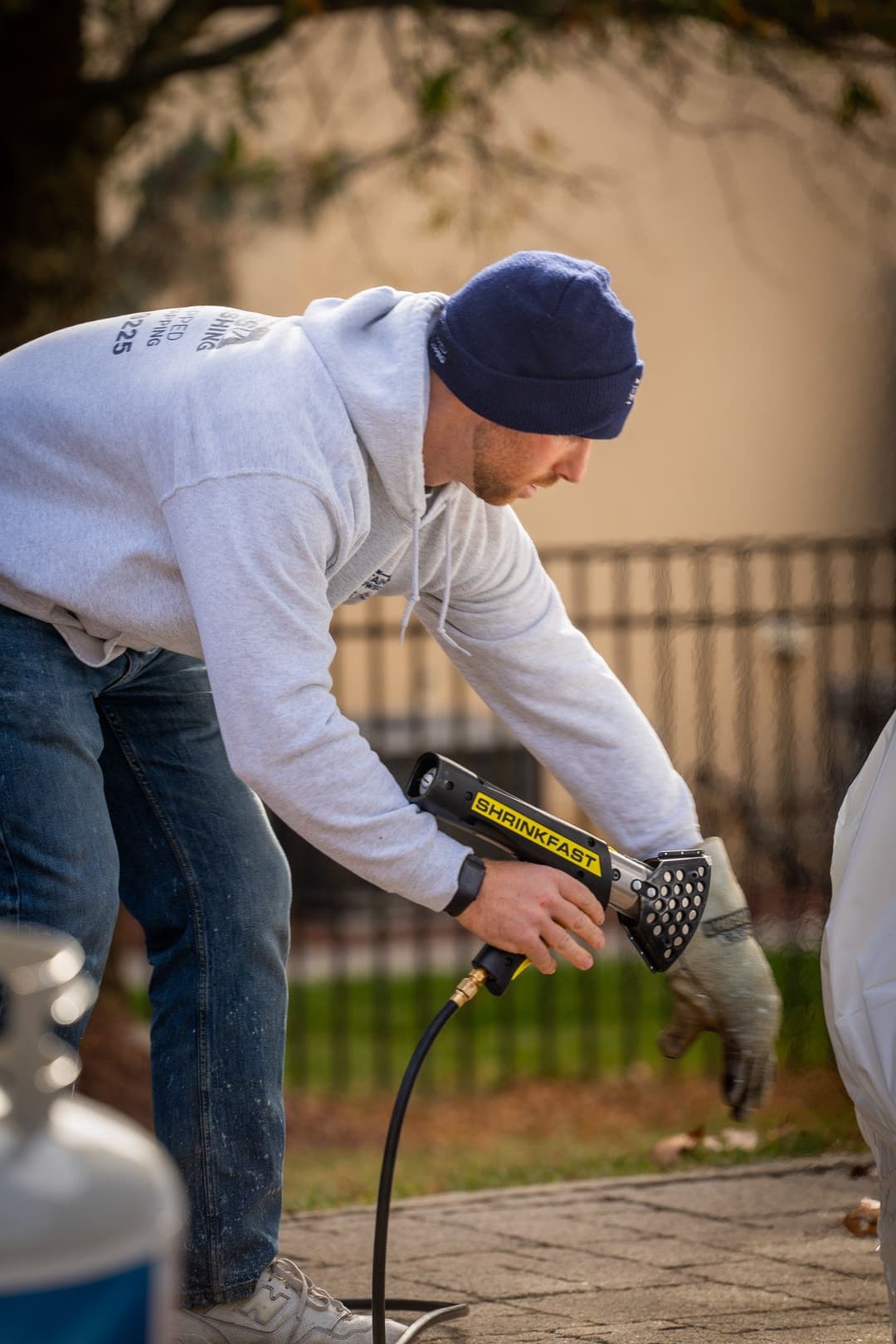 Man using a heat gun to remove paint from a surface outdoors, wearing a gray hoodie and hat.
