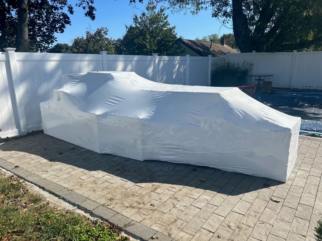 Car covered with a protective white cover parked on a paved driveway beside a white fence.