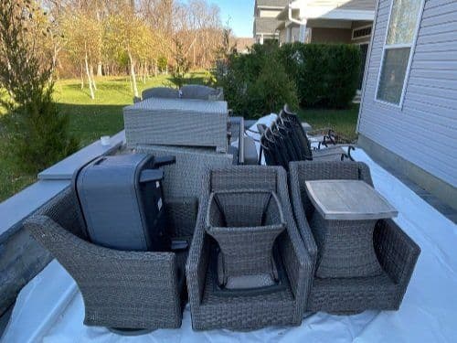 Stacked outdoor furniture including chairs, tables, and a grill on a white tarp in a yard.