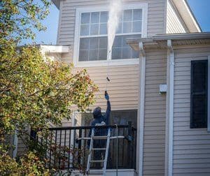Person using a pressure washer on a home's exterior, cleaning upper windows and siding.