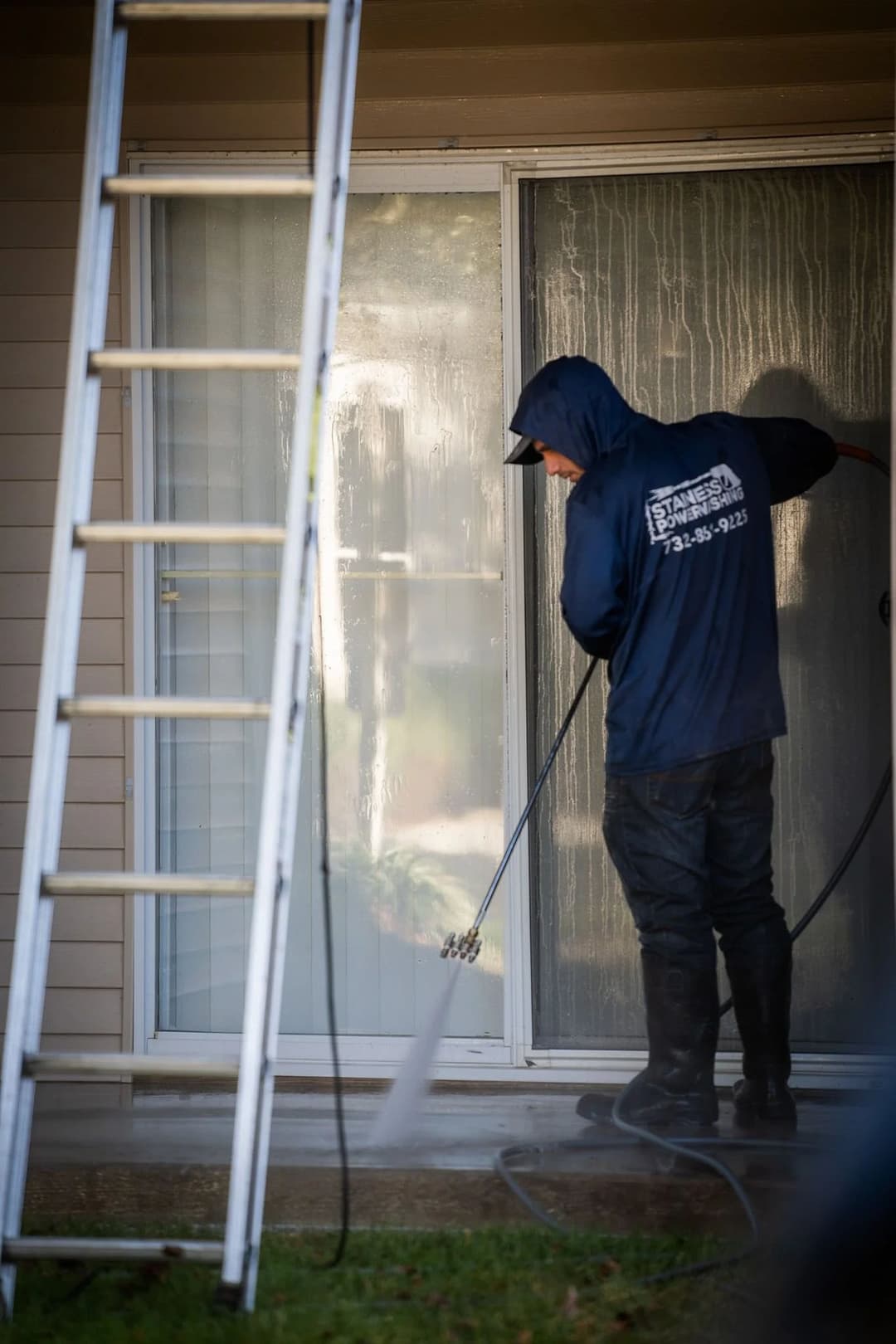 Person using a pressure washer to clean a glass door, with a ladder in the background.