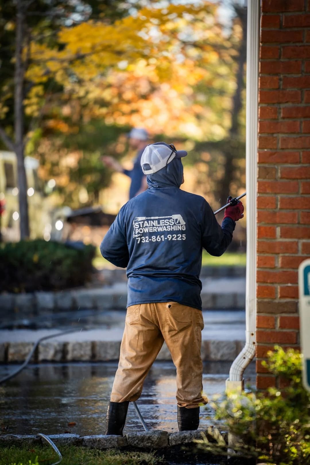 Power washer in action, cleaning a brick exterior with trees and driveway in the background.
