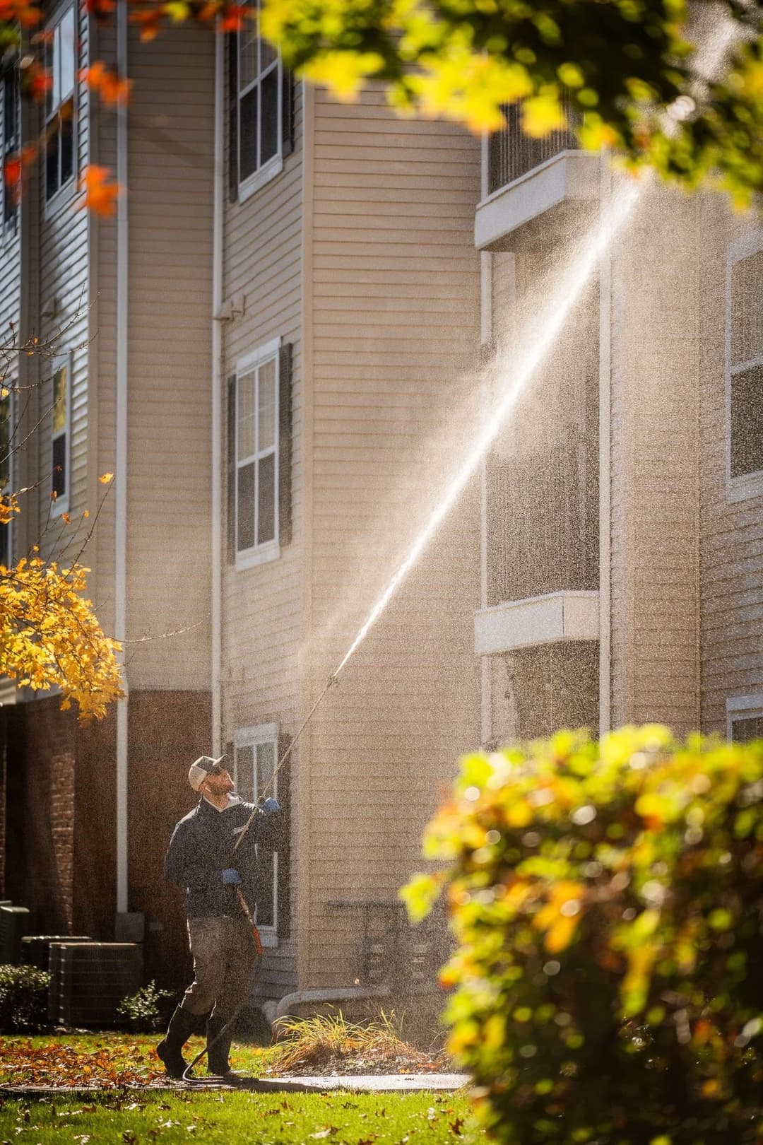 Man using a pressure washer on an apartment building exterior in bright sunlight.