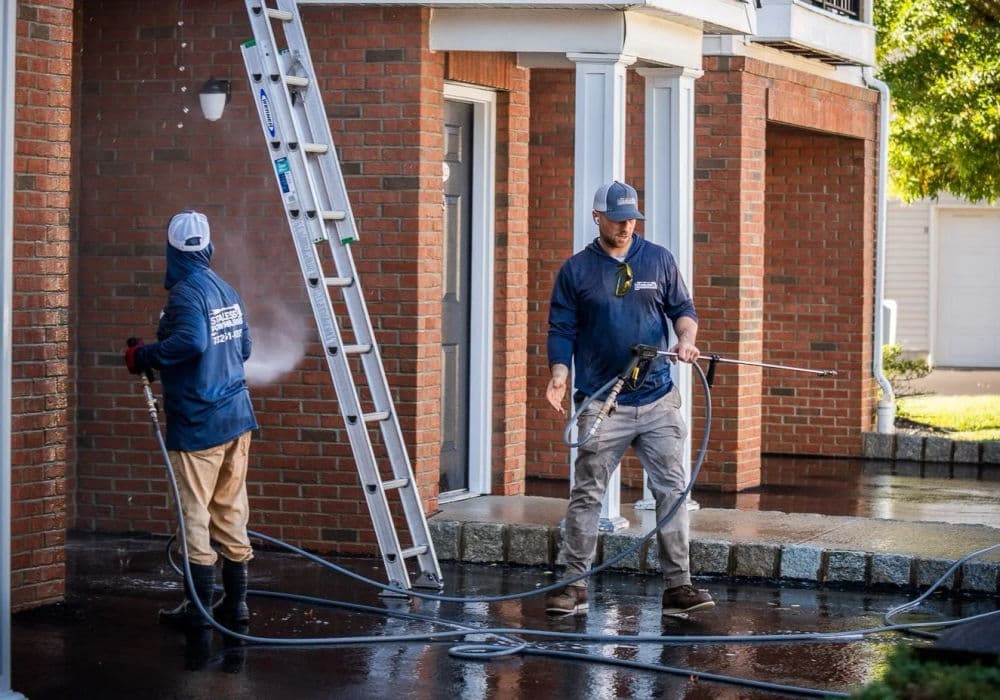 Two professionals pressure washing a brick building using hoses and equipment.